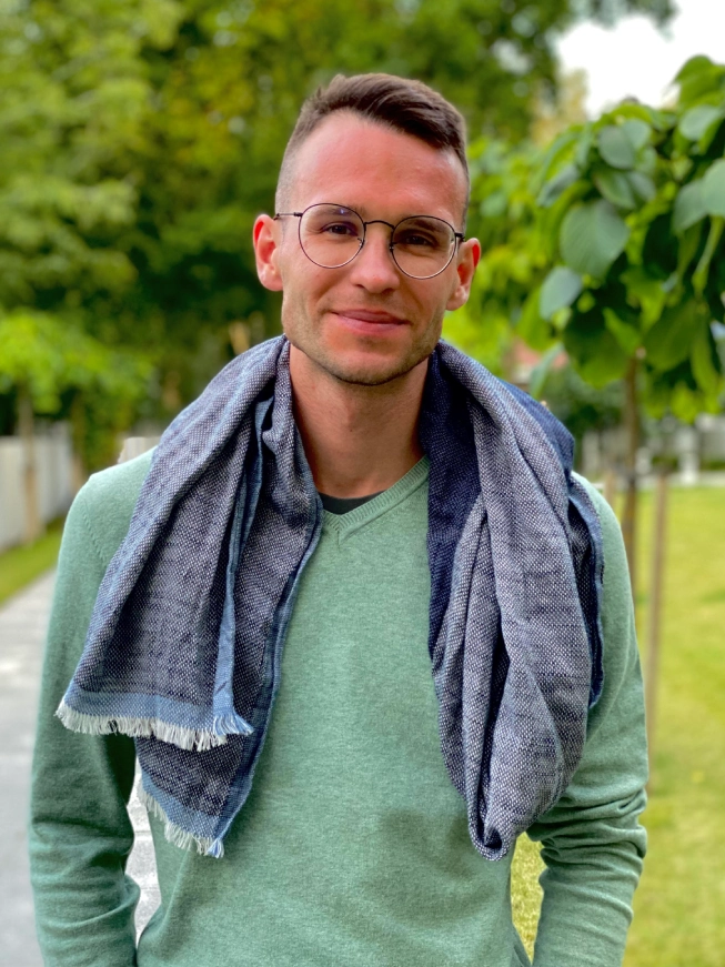 Mężczyzna w okularach i zielonym swetrze stoi na zewnątrz, z szalikiem na ramionach.