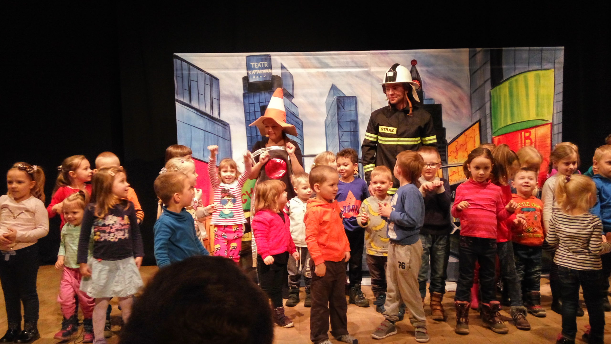 Dzieci stoją na scenie z aktorem przebranym za strażaka i aktorką w czapce drogowej.