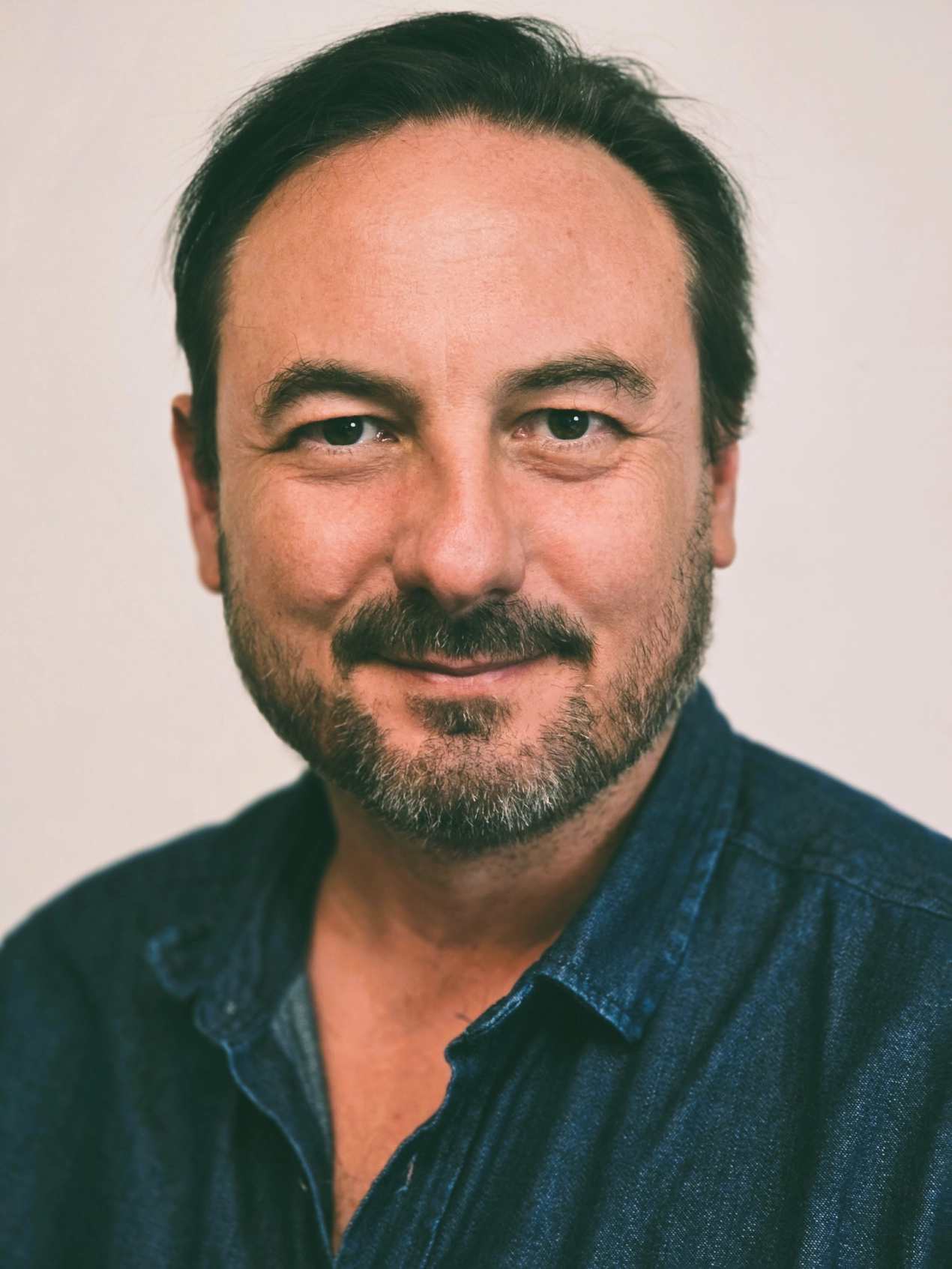 Mężczyzna z brodą w niebieskiej koszuli patrzy z uśmiechem prosto w obiektyw.