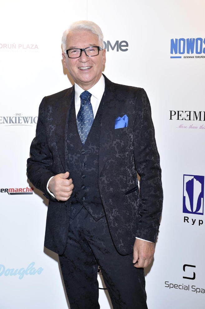 Mężczyzna w garniturze pozuje na tle ścianki z logotypami podczas wydarzenia.