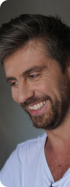 Uśmiechający się mężczyzna z brodą w białej koszulce, patrzący w dół.