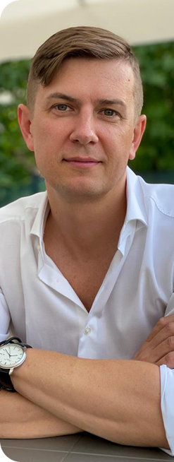 Mężczyzna w białej koszuli siedzi przy stole na zewnątrz, patrzy prosto w obiektyw.