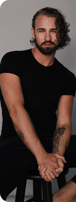 Mężczyzna w czarnym t-shircie siedzi na stołku, splatając dłonie, patrzy w obiektyw.